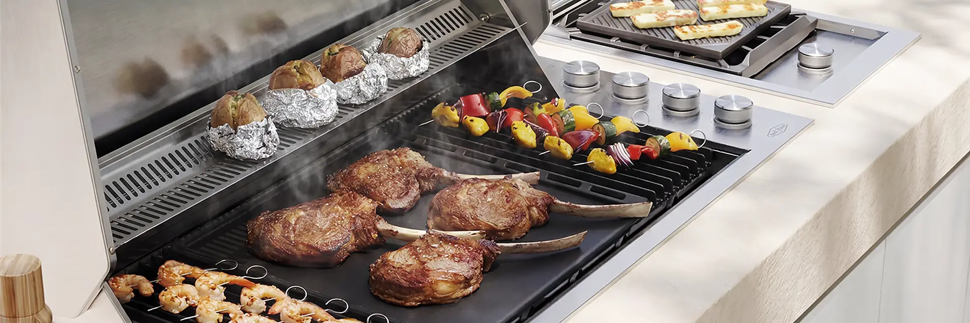 BeefEater ProLine built-in barbecue with its hood open, cooking steaks, prawns and vegetable skewers, with halloumi on the side burner beside it