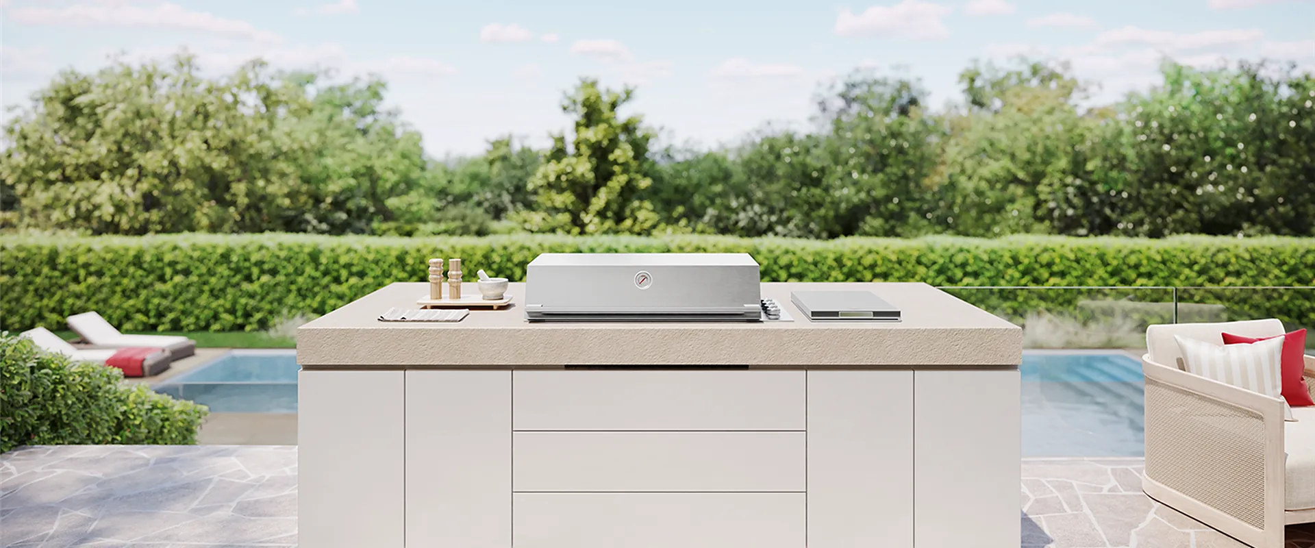 BeefEater ProLine built-in barbecue in stainless steel set into a white outdoor kitchen island on a stone terrace, with a pool and a clipped hedge behind