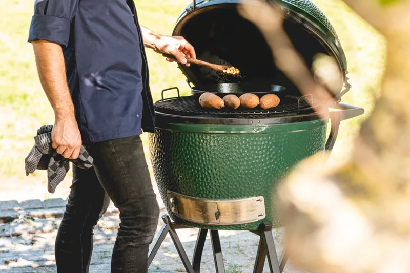 Cook in a navy jacket turning sweet potatoes on the grate of an open Big Green Egg 2XL kamado outdoors