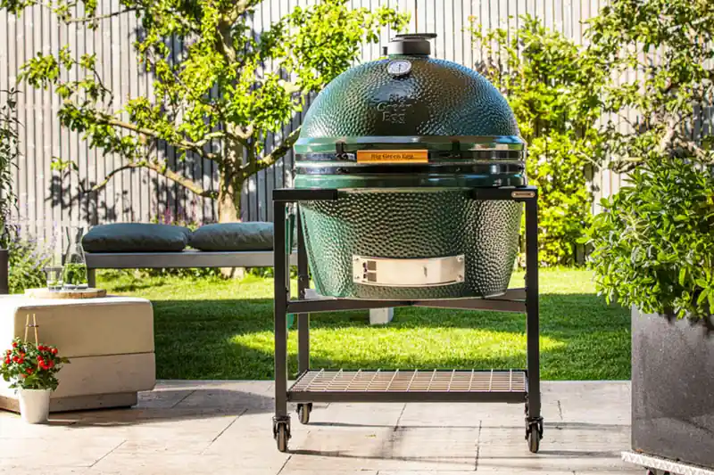 Big Green Egg 2XL kamado in a black modular frame on wheels on a stone patio, with a lawn and a fruit tree behind