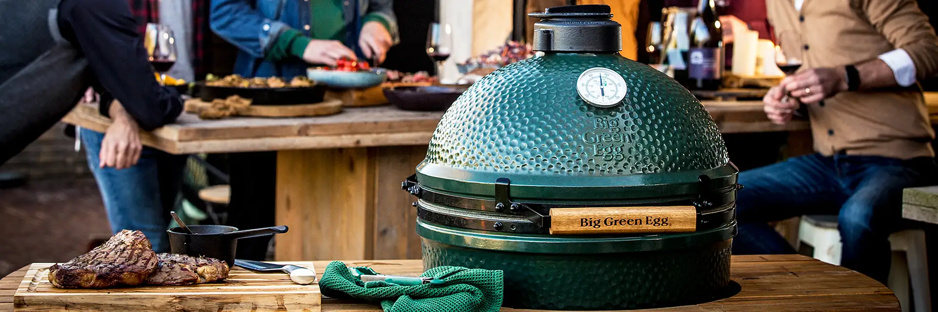 Big Green Egg kamado set into a wooden outdoor kitchen counter with grilled steaks on a board, while a group of friends share food and wine behind it at dusk