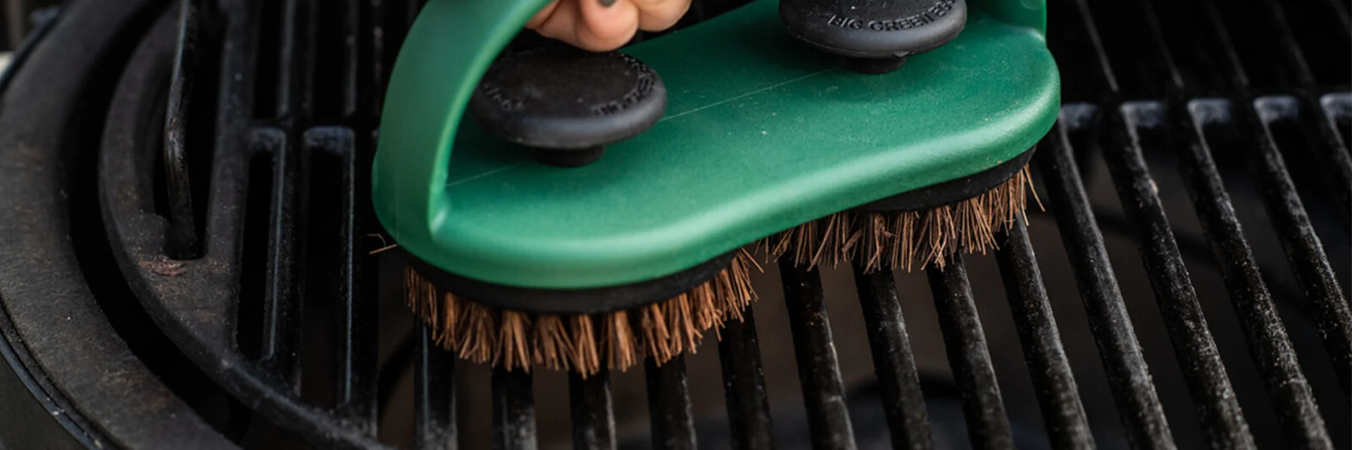 Hand scrubbing the cast-iron cooking grid of a Big Green Egg kamado with a green-handled brush