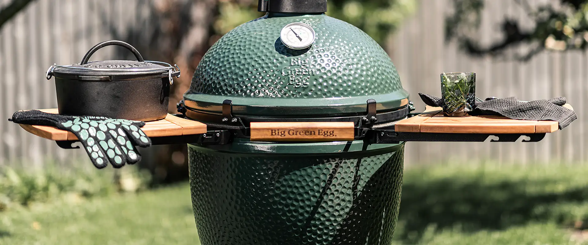 Big Green Egg kamado with acacia wood side shelves and a cast-iron pot, in front of a garden fence