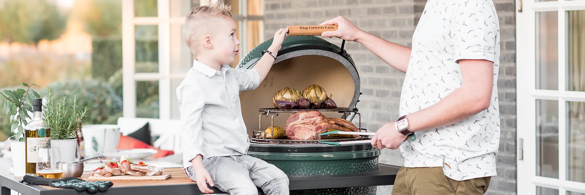 Father and son opening a Big Green Egg kamado built into an outdoor table, with roast meat and artichokes on the grid, on a brick-walled terrace