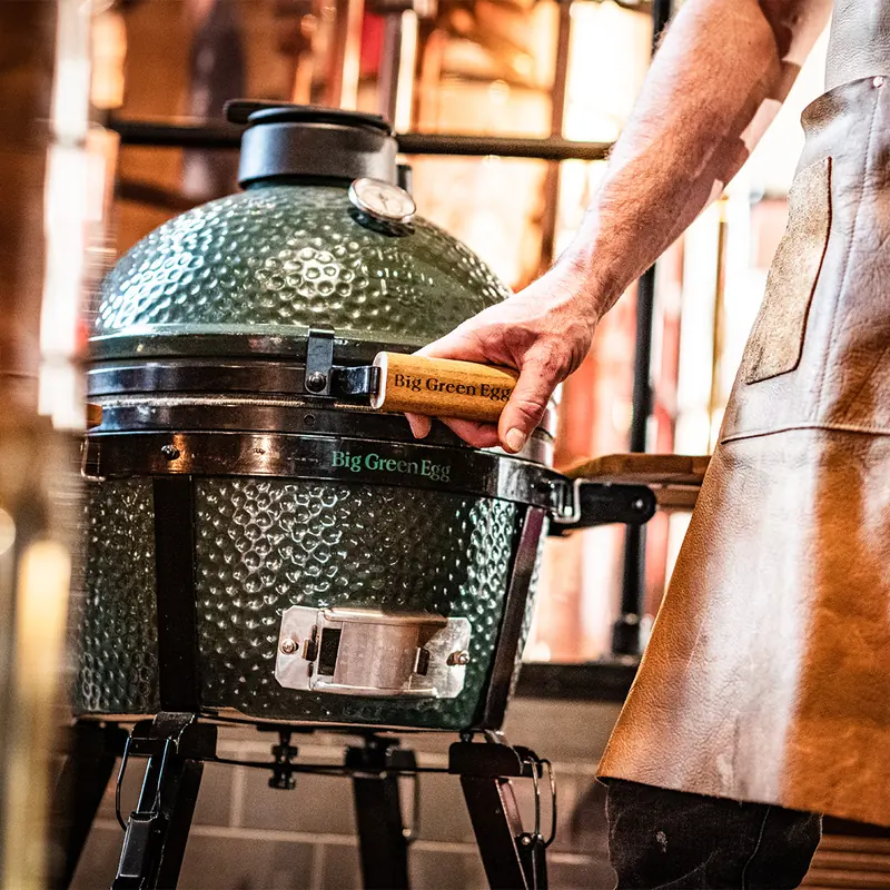 Cook in a leather apron lifting the lid of a Big Green Egg kamado by its wooden handle