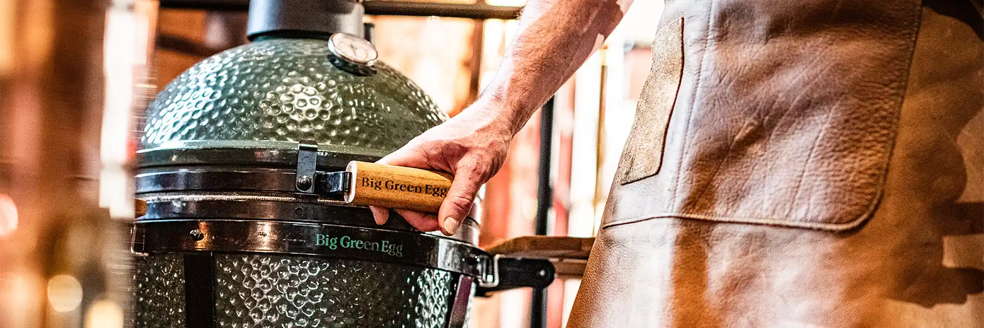 Cook in a leather apron lifting the lid of a Big Green Egg kamado by its wooden handle