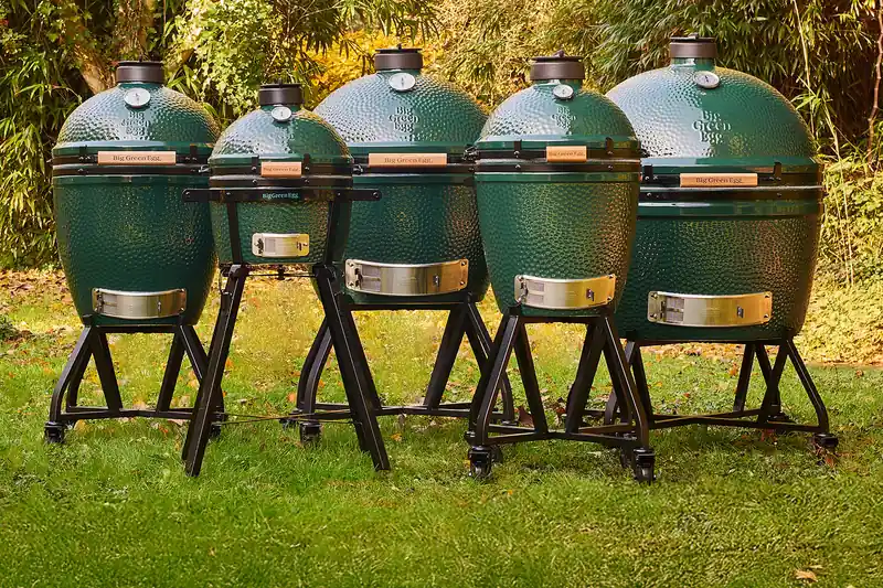 Five Big Green Egg kamado grills in different sizes lined up on a garden lawn with autumn foliage behind