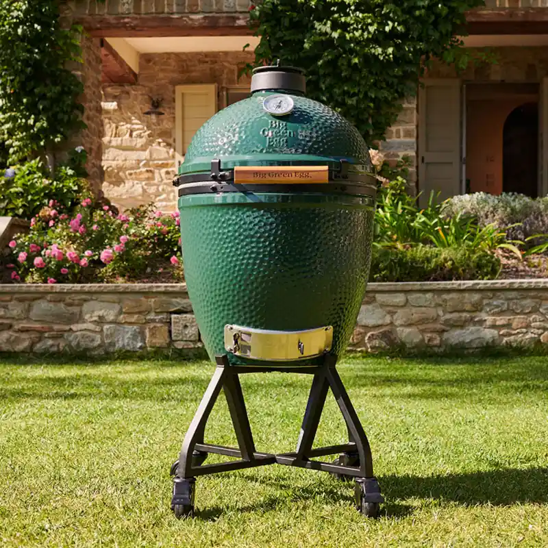 Big Green Egg ceramic kamado on its rolling nest on a lawn, in front of a stone house with climbing plants and pink flowers