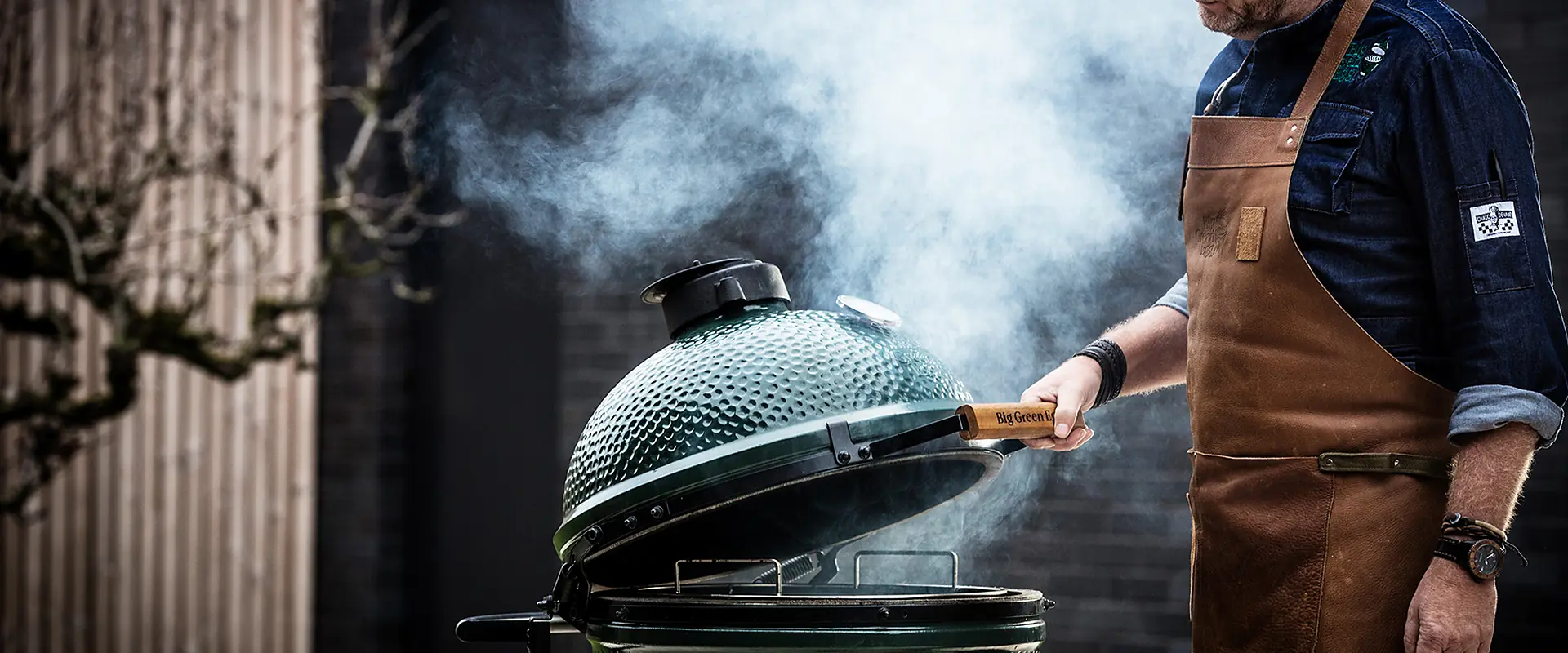 Chef in a leather apron opening a smoking Big Green Egg kamado in front of a dark timber wall
