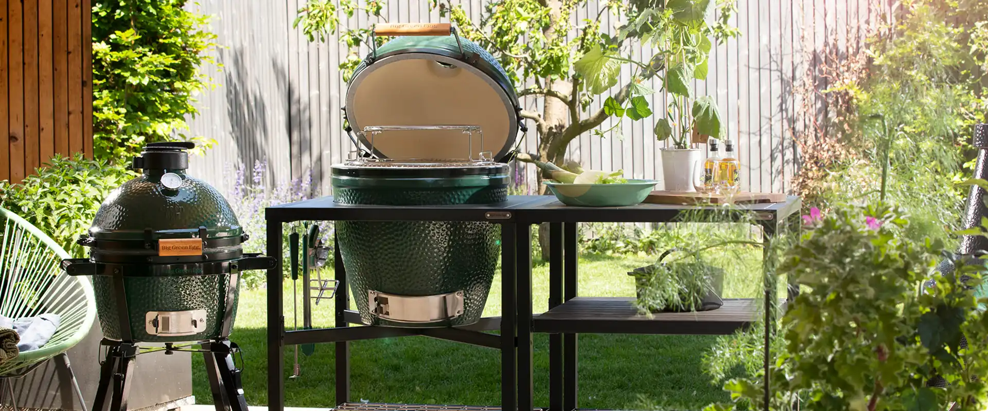 Big Green Egg MiniMax on a portable stand beside a larger Egg with its lid open in a black modular table, on a sunny garden patio