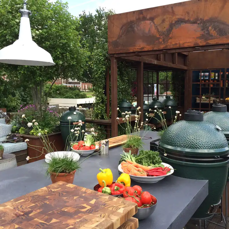 Big Green Egg kamados beside a concrete outdoor kitchen island laid with vegetables, chopping boards and a pendant lamp in a garden