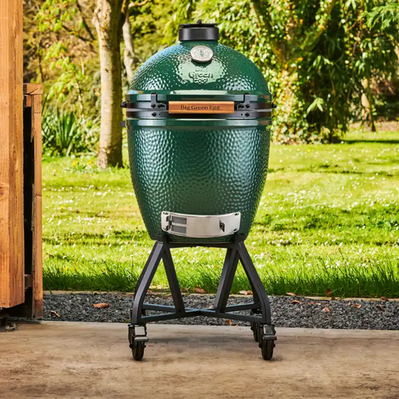 Big Green Egg Large kamado on a rolling stand on a concrete terrace, with a sunny lawn and trees behind