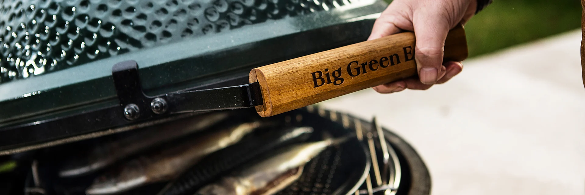 Hand lifting the lid of a Big Green Egg kamado by its wooden handle, with whole fish cooking on the grid inside