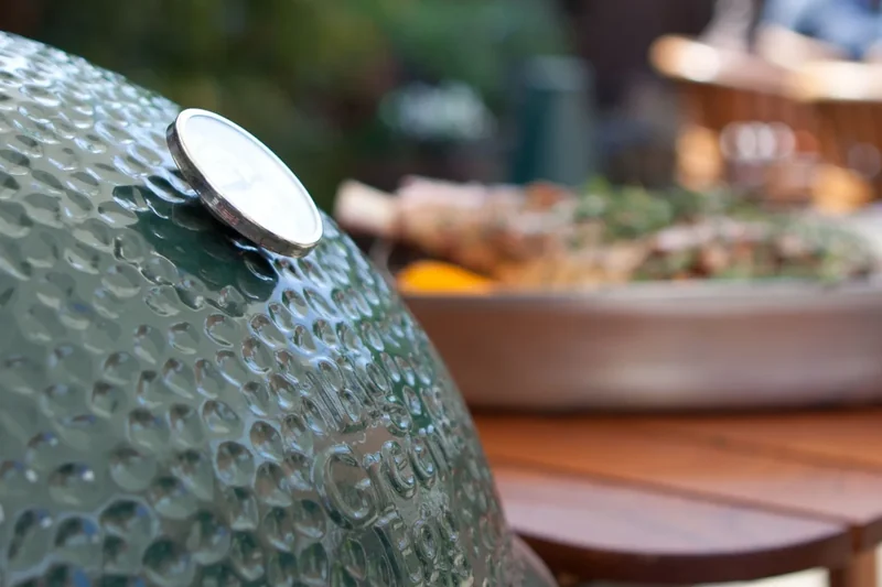 Big Green Egg Medium kamado beside a timber garden table with a roasting dish, guests chatting in the background