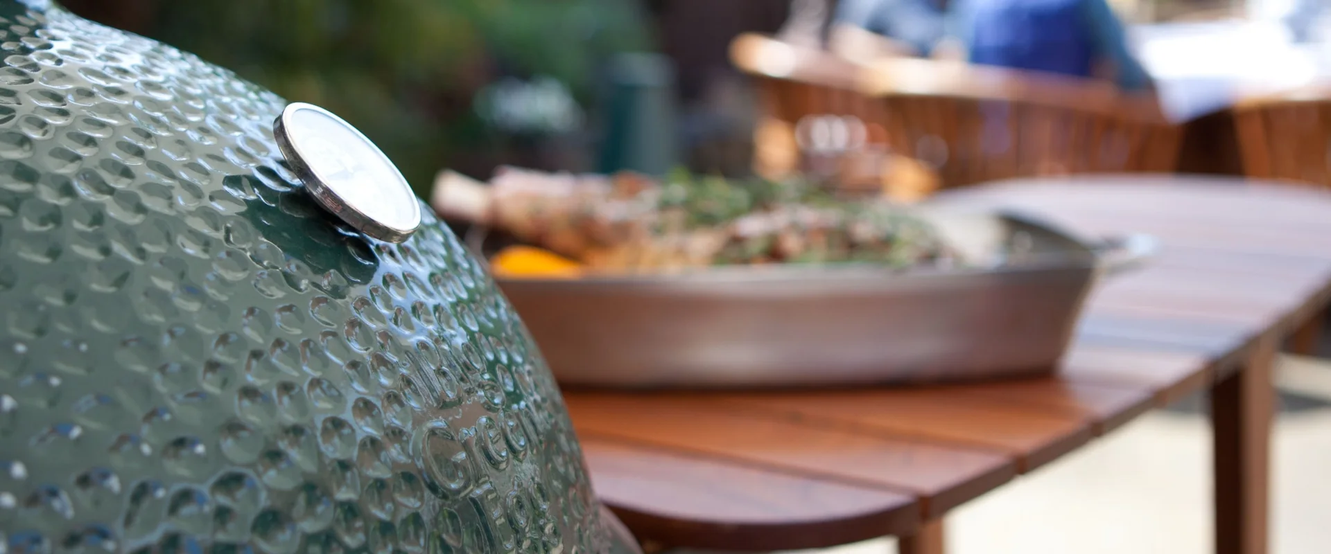 Big Green Egg Medium kamado beside a timber garden table with a roasting dish, guests chatting in the background