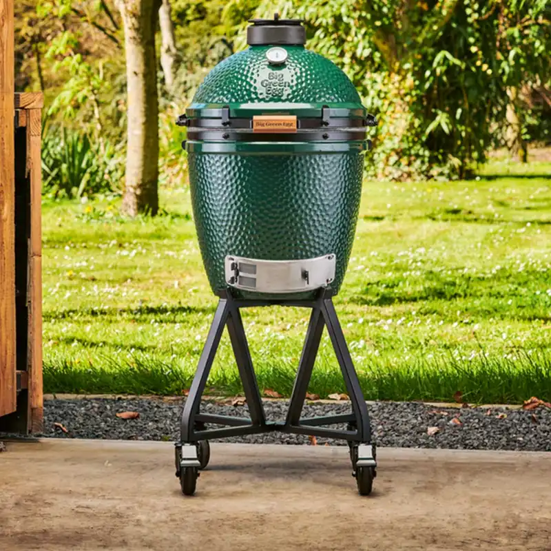 Big Green Egg Medium kamado on a rolling stand on a concrete terrace, with a sunny lawn and trees behind