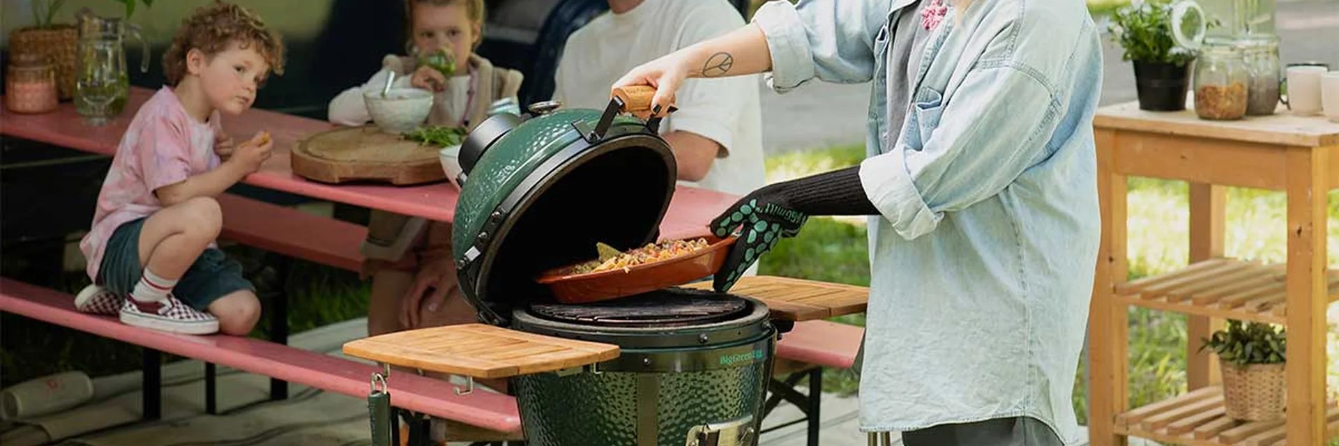 A cook lifting a dish from an open Big Green Egg MiniMax while children wait at a garden lunch table