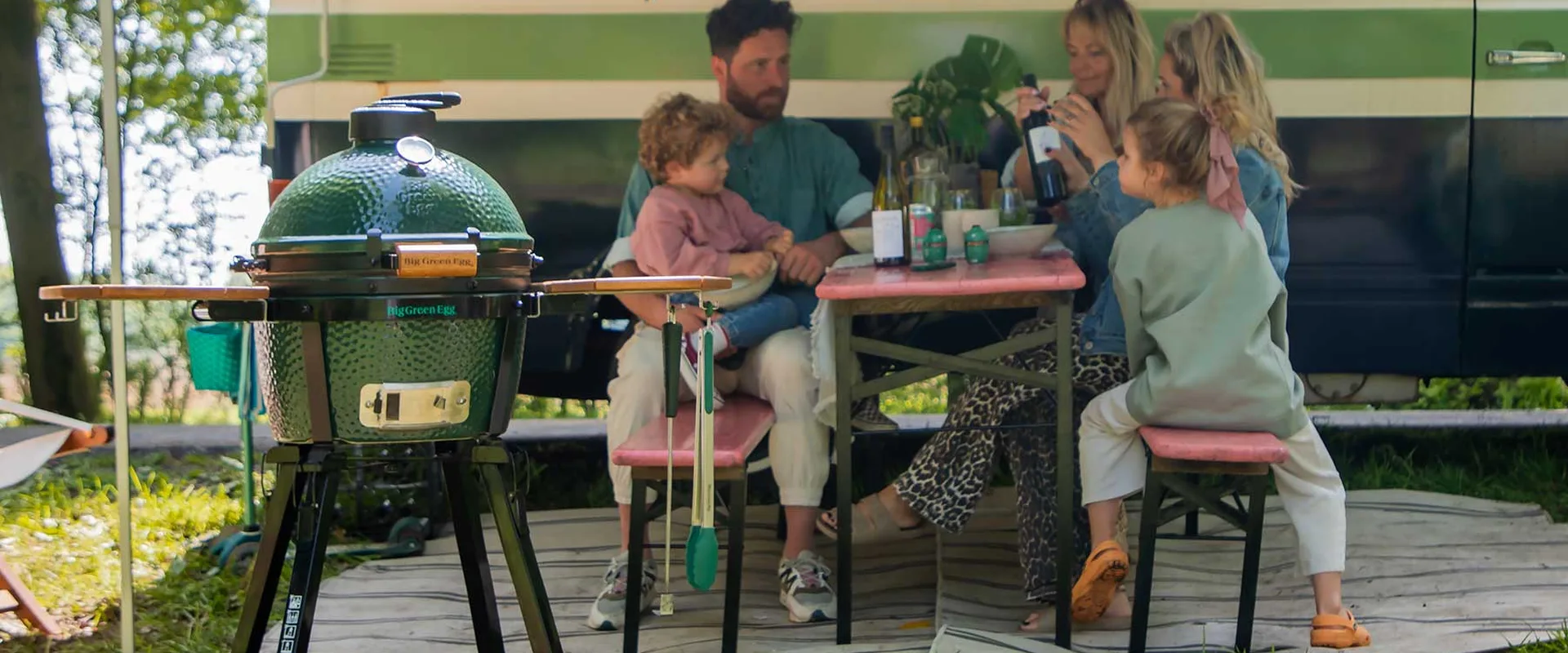 A family having lunch at a garden table beside a Big Green Egg MiniMax on its stand