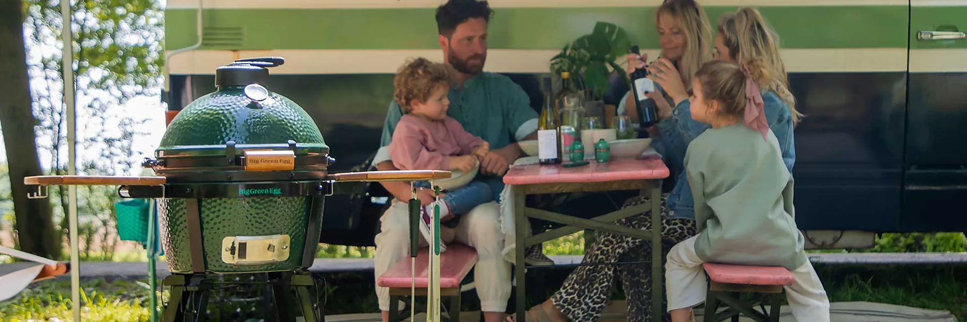 Big Green Egg MiniMax on its stand on the grass, beside a family eating at a table outdoors