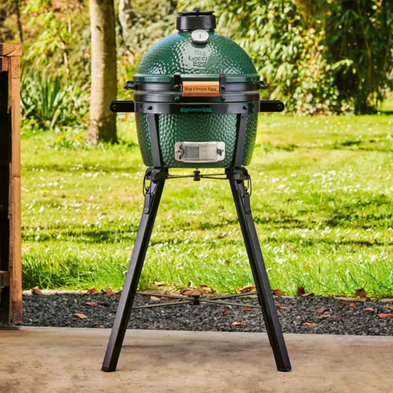 Big Green Egg MiniMax kamado on its tall portable stand on a concrete terrace, with a sunny lawn behind