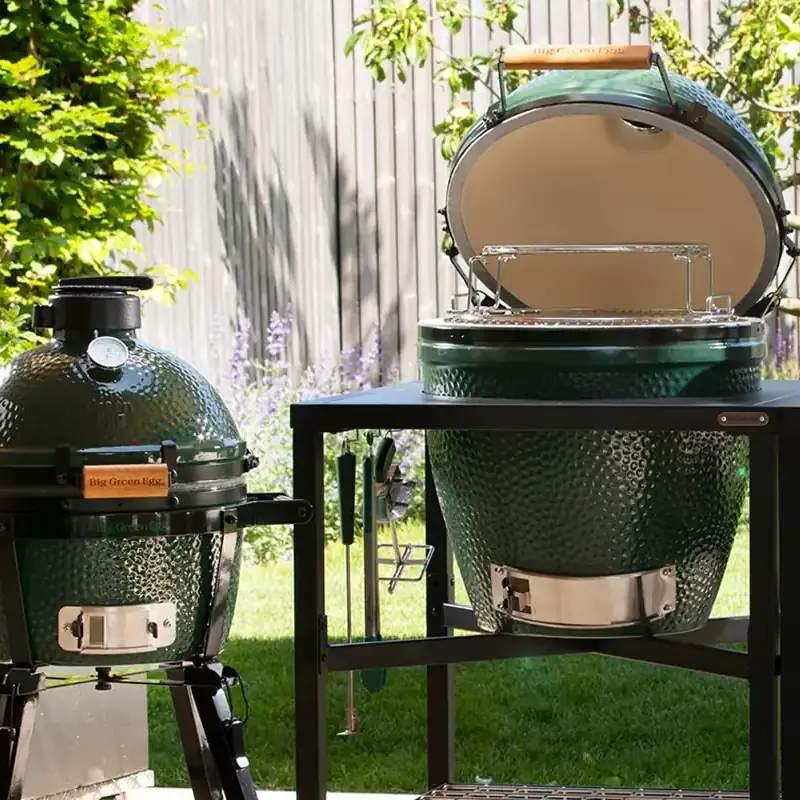 Big Green Egg MiniMax on its carrier beside a Large EGG with the lid open, set into a black steel table in a sunny garden