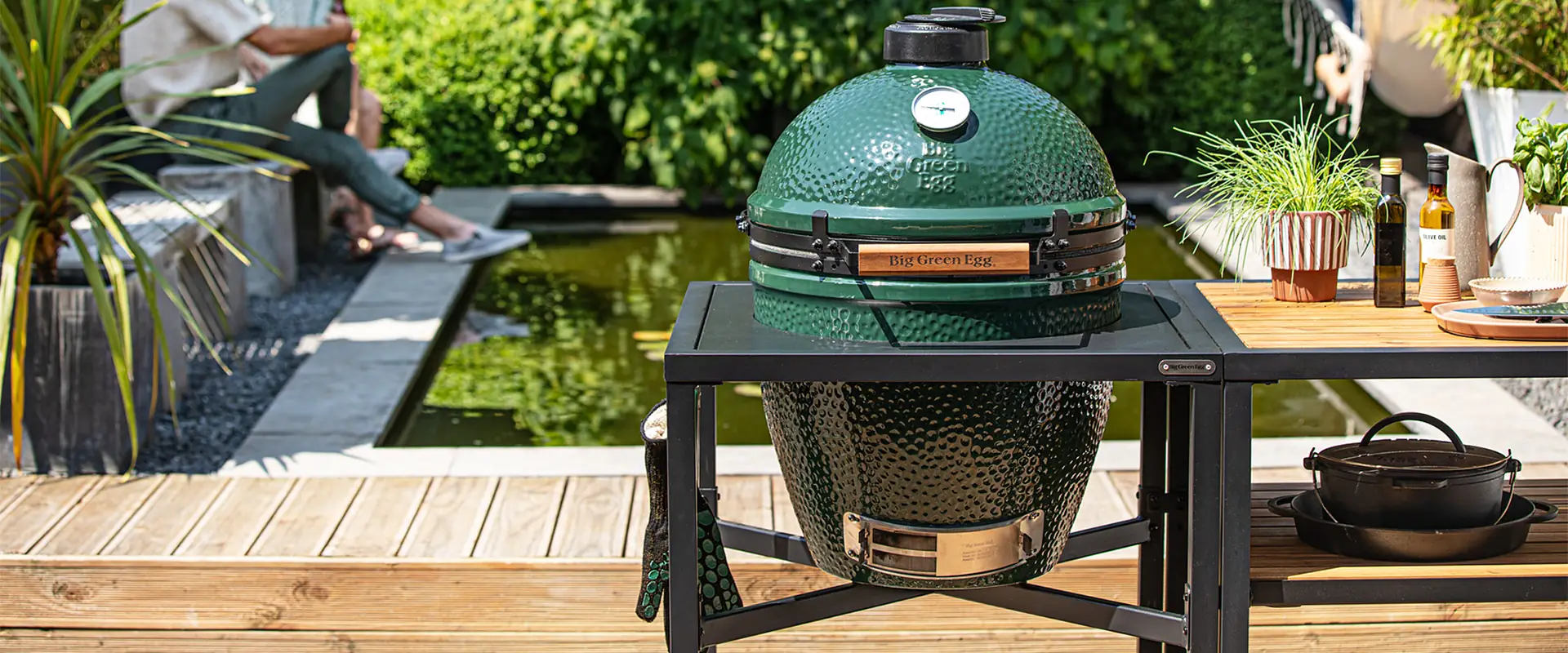 Big Green Egg kamado in a black Modular EGG Frame with an acacia worktop beside it, on a timber deck by a garden pond, with two people sitting in the background