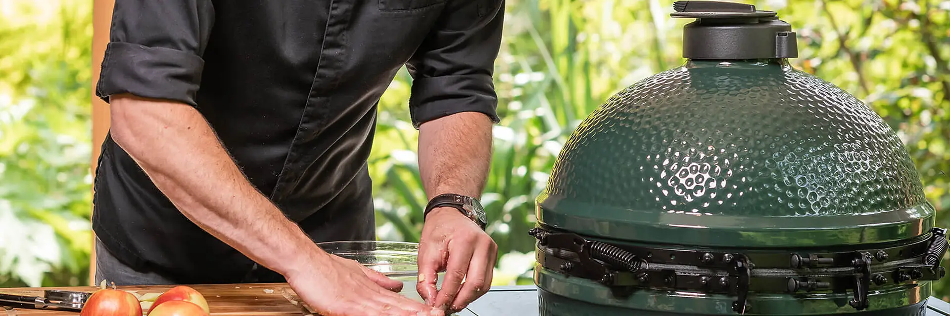 Chef in a black jacket pressing pastry into a small dish on a wooden worktop beside a Big Green Egg kamado set into the counter, in a leafy garden
