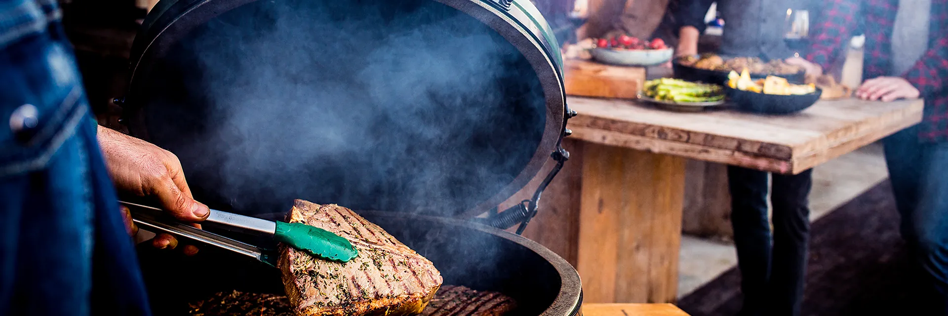 Steak being turned with tongs on a Big Green Egg with the lid open, with guests around a table behind