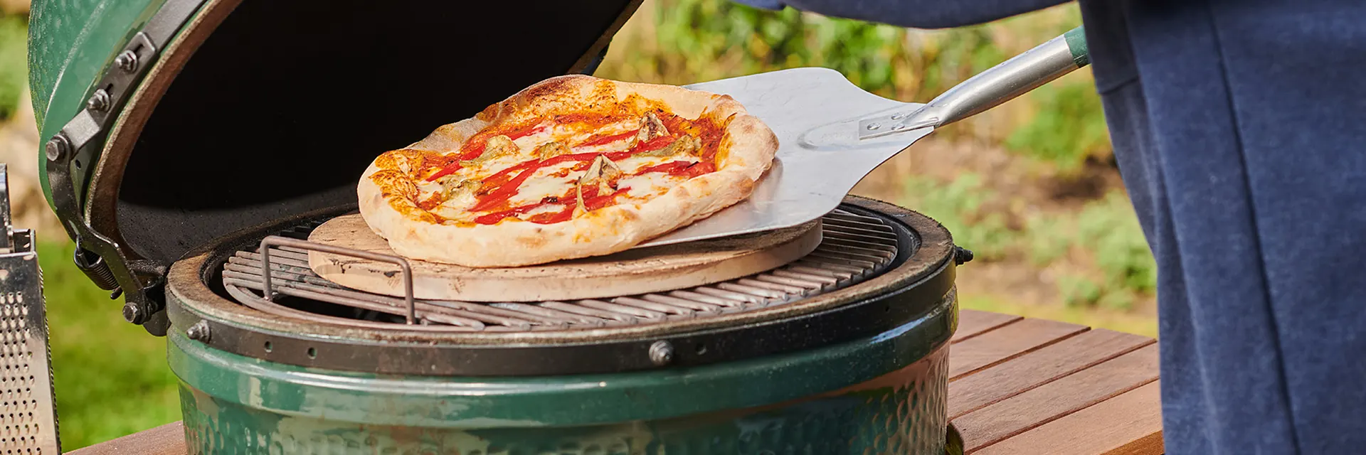 Pizza being lifted with a peel from the baking stone of a Big Green Egg kamado set in a slatted wooden table in a garden
