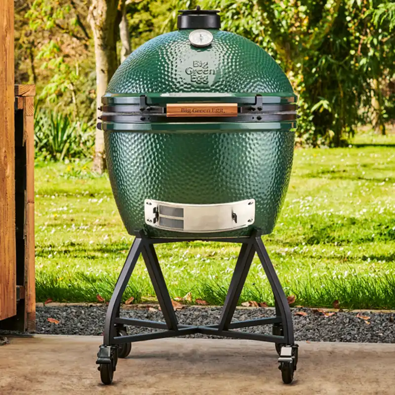 Big Green Egg XLarge kamado on a rolling stand on a concrete terrace, with a sunny lawn and trees behind