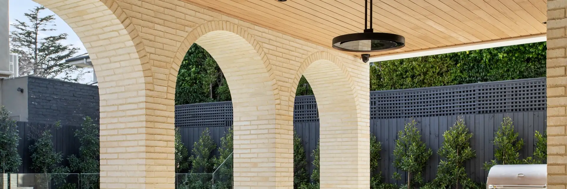 Bromic Eclipse Electric pendant heater hanging from a timber ceiling above a dining table, framed by pale brick arches and a garden