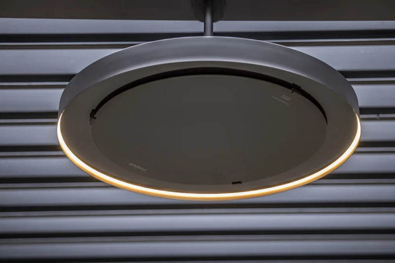 Close-up from below of a Bromic Eclipse pendant heater with its LED light ring glowing, against a dark louvred pergola roof