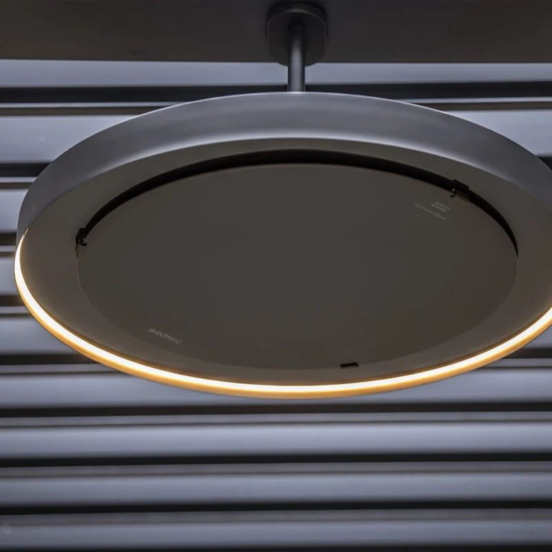 Close-up from below of a Bromic Eclipse pendant heater with its LED light ring glowing, against a dark louvred pergola roof