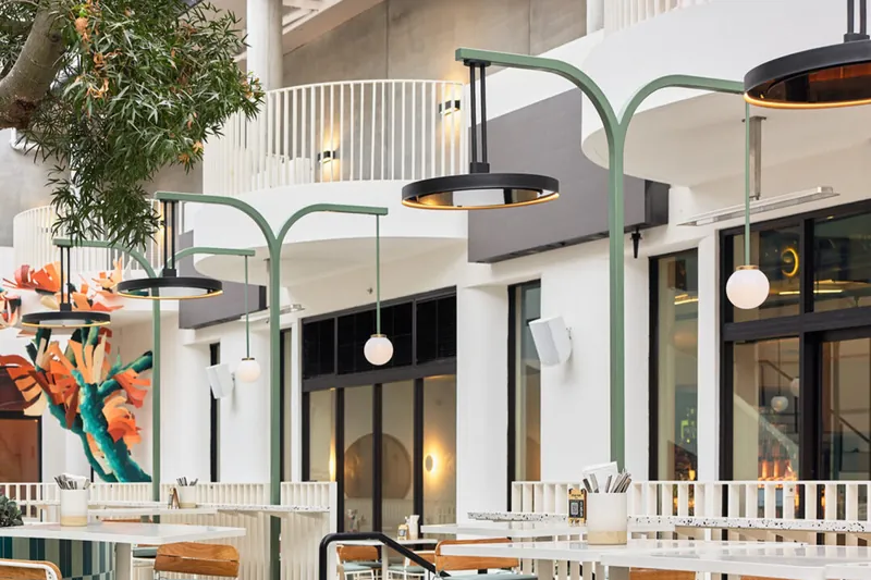 Bromic Eclipse pendant heaters hanging from sage-green arched posts over a restaurant courtyard beside a large tree