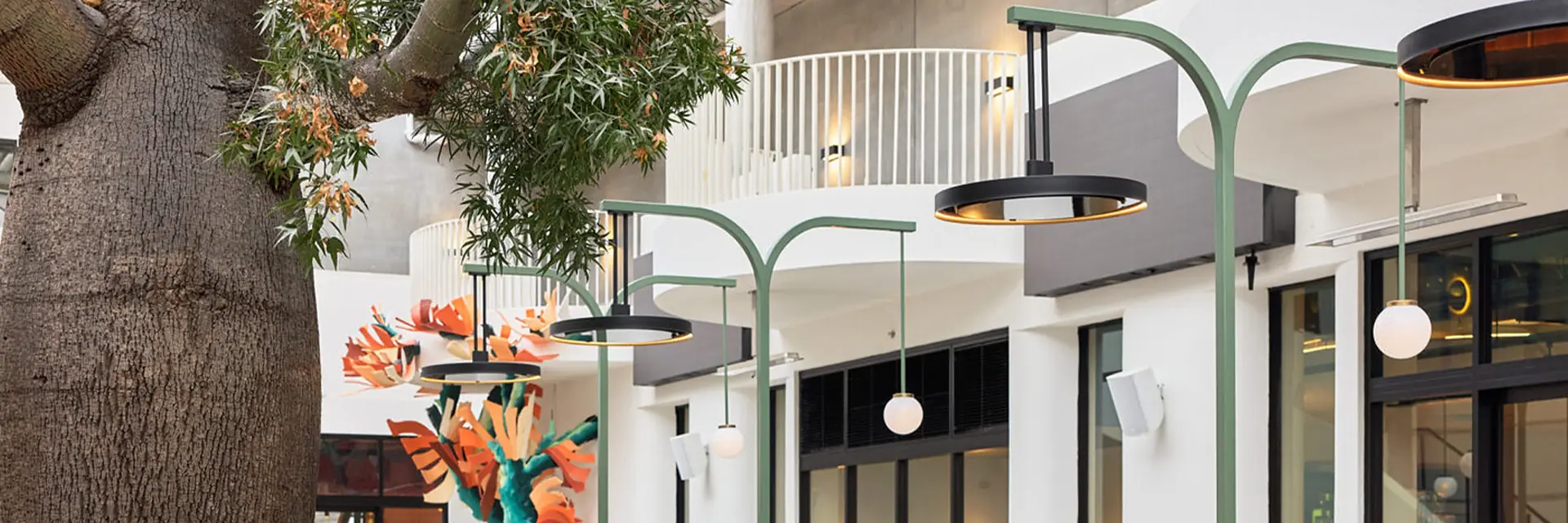 Bromic Eclipse pendant heaters hanging from sage-green arched posts over a restaurant courtyard beside a large tree