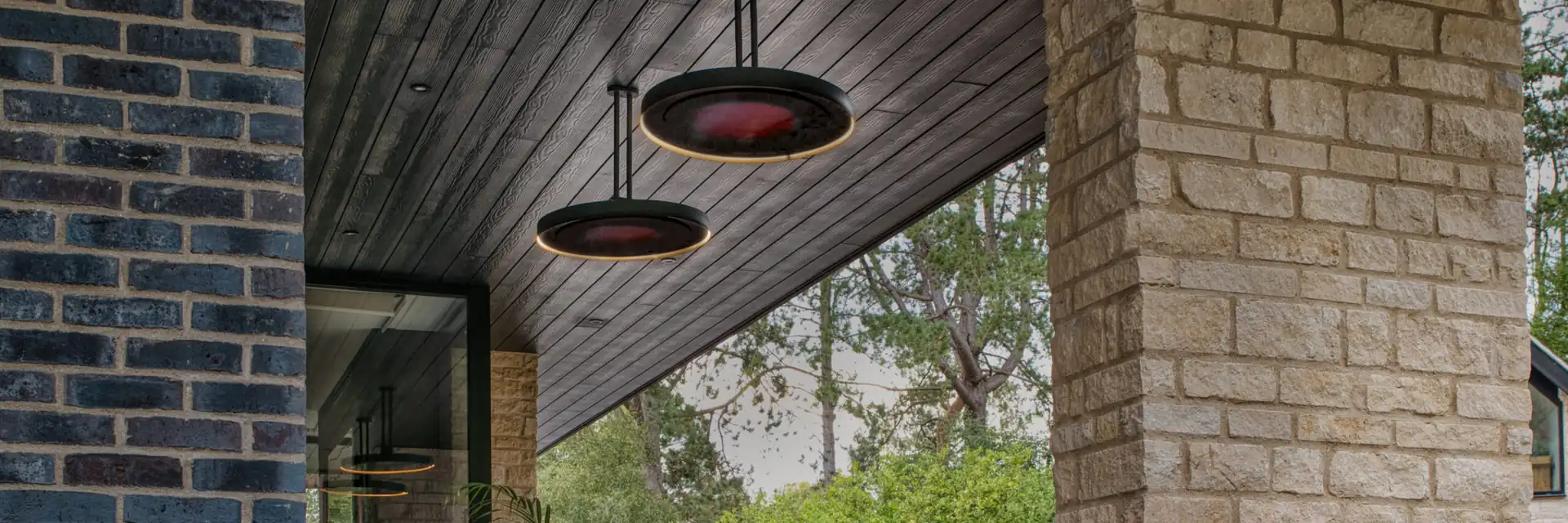 Two Bromic Eclipse Electric pendant heaters glowing beneath a dark timber soffit between brick and stone pillars, above a garden seating area