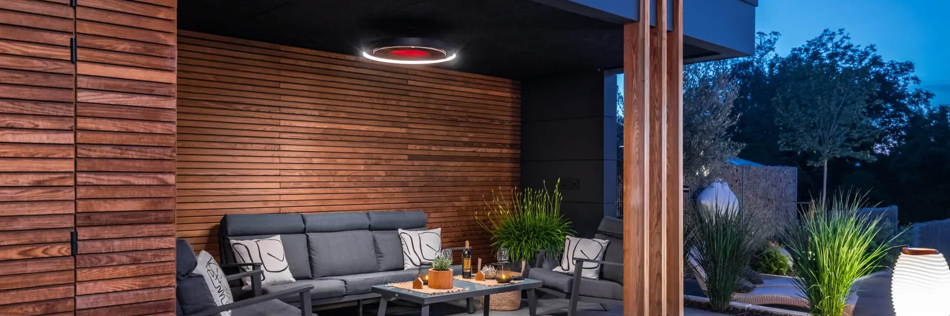 Bromic Eclipse Electric pendant heater with its light ring on above an outdoor sofa, set against a timber-slat wall at dusk