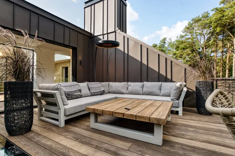 Bromic Eclipse Smart-Heat Portable heater beside a grey corner sofa and a timber coffee table on a roof deck, with pine trees behind