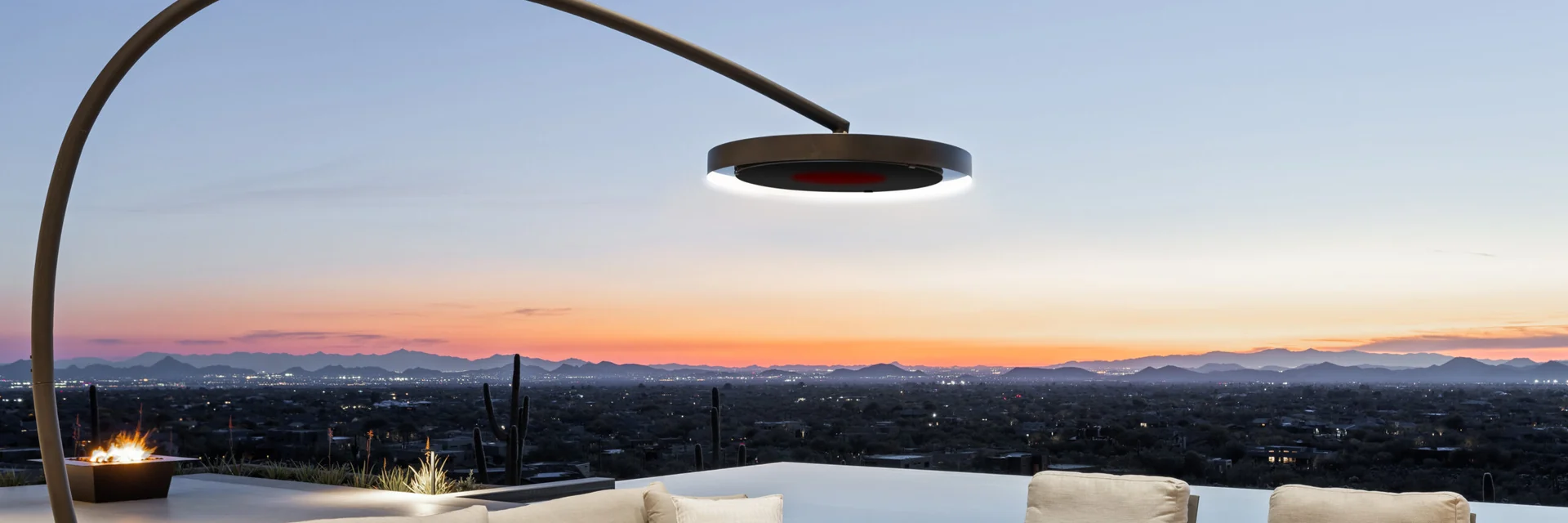 Bromic Eclipse Portable heater arching over cream sofas on a terrace beside a pool, overlooking a desert valley at sunset