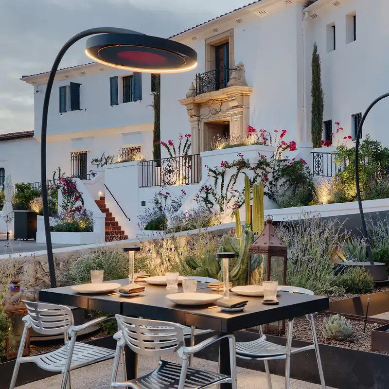 Two Bromic Eclipse Portable heaters arching over set dining tables at dusk, in front of a white Spanish-style mansion with cypresses and bougainvillea