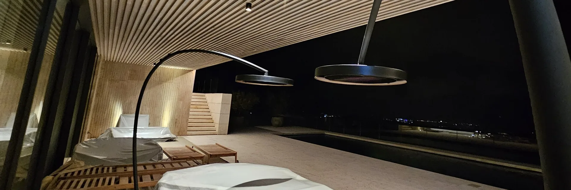Two Bromic Eclipse portable heaters with arched stands over the loungers of a villa terrace in Sotogrande at night, under a slatted timber ceiling
