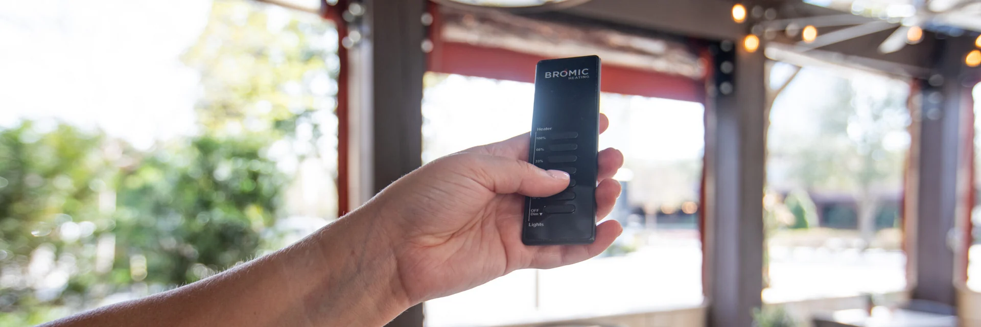 Hand holding a Bromic remote control in front of a restaurant terrace with round tables