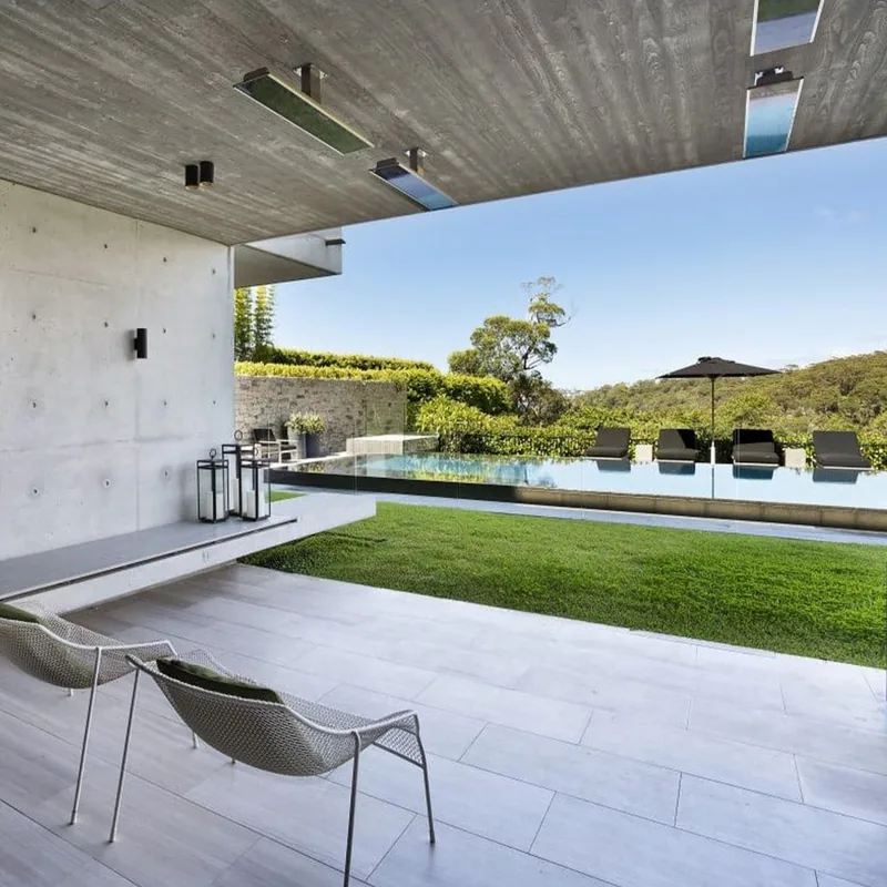 Bromic electric heaters suspended from a concrete ceiling over a terrace with lounge chairs, a lawn and an infinity pool beyond
