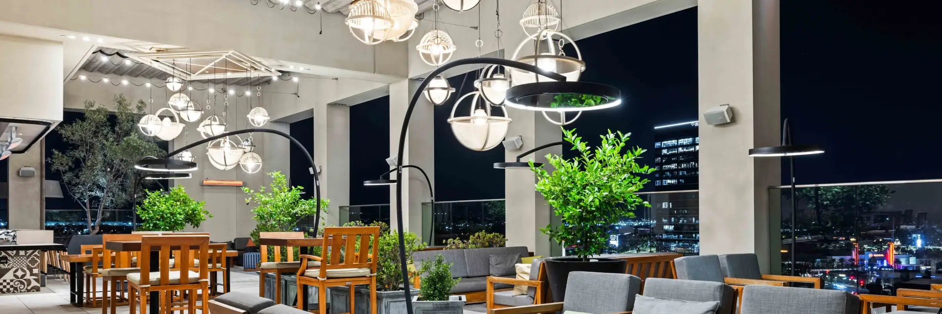 Bromic heaters on curved poles between the tables and sofas of a rooftop lounge at night, under globe pendant lights