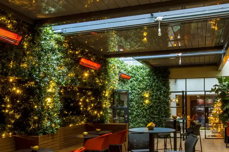 Bromic electric heaters glowing on the walls of a covered restaurant terrace with a living green wall, fairy lights, red armchairs and a timber deck