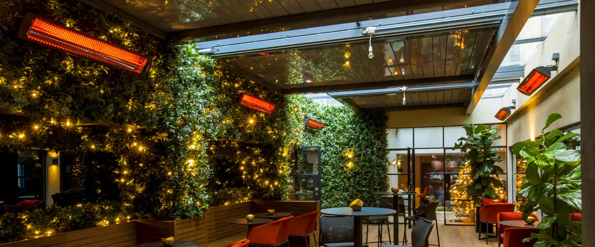 Bromic electric heaters glowing on the walls of a covered restaurant terrace with a living green wall, fairy lights, red armchairs and a timber deck