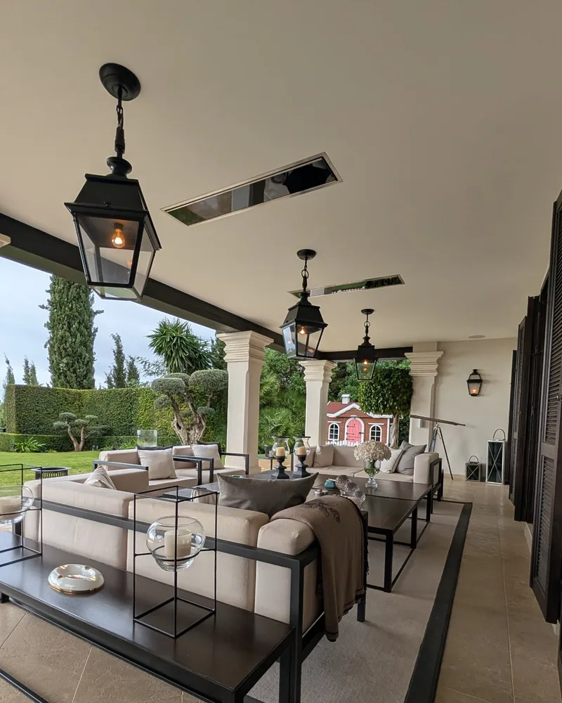 Black Bromic Platinum Electric heaters recessed in the ceiling of a covered villa terrace, with hanging black lanterns, cream outdoor sofas and a garden beyond