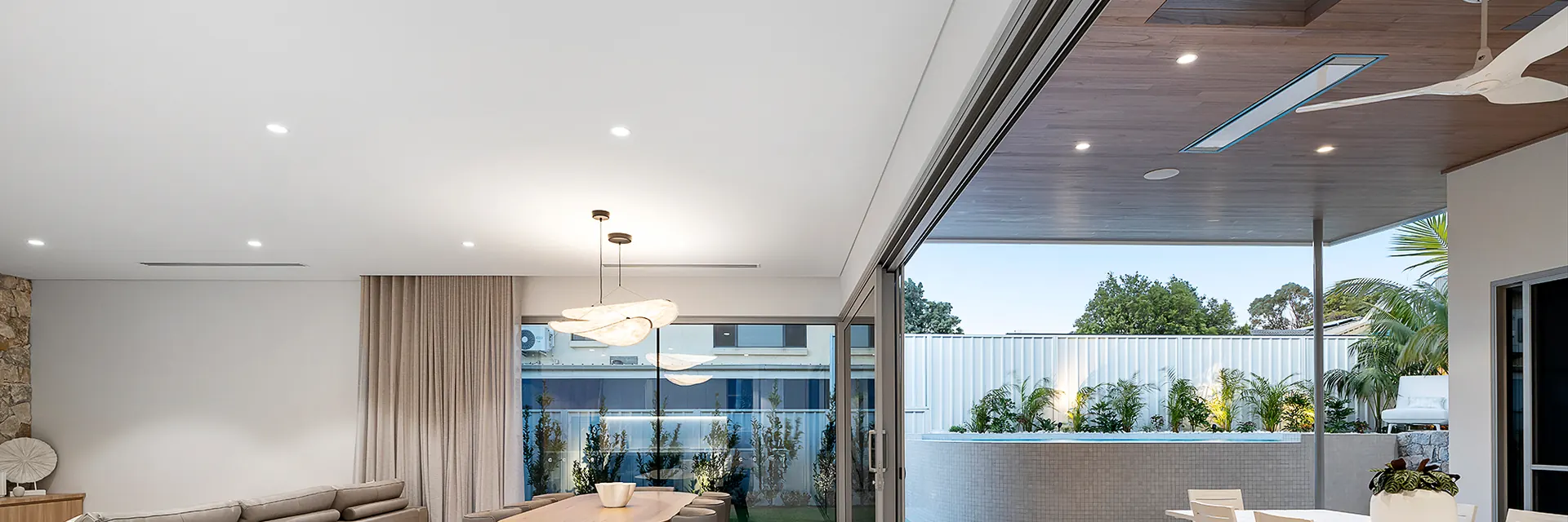Bromic Platinum Electric heater recessed in a timber ceiling beside a fan, above an alfresco dining table that opens to a bright living room