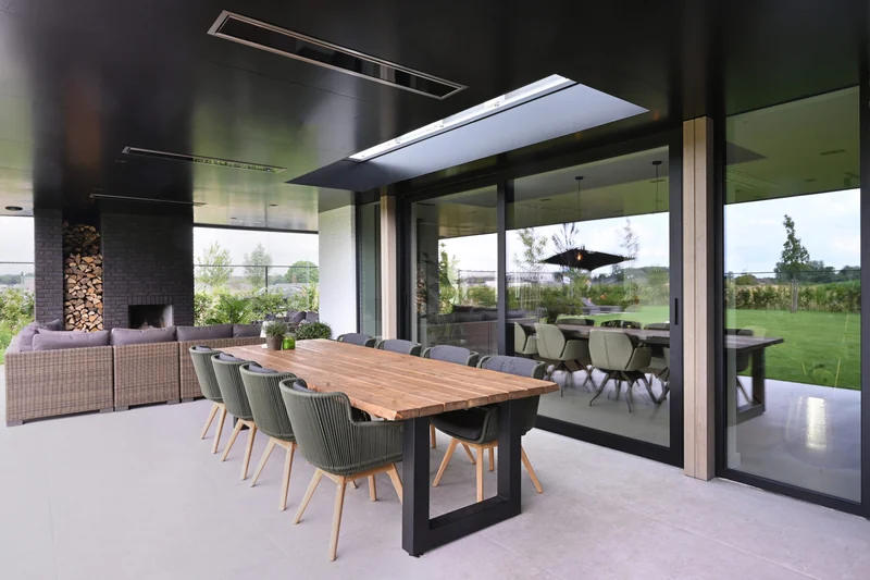 Black Bromic Platinum Electric heaters recessed in the dark ceiling of a covered terrace with a long wooden dining table and garden views