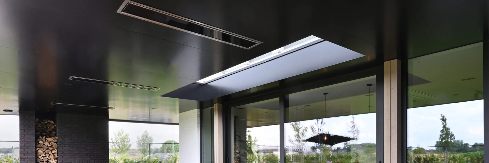 Black Bromic Platinum Electric heaters recessed in the dark ceiling of a covered terrace with a long wooden dining table and garden views